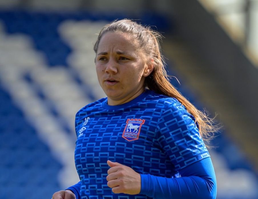 Female soccer player in a blue training kit jogging on the field stadium seating in the blurred background