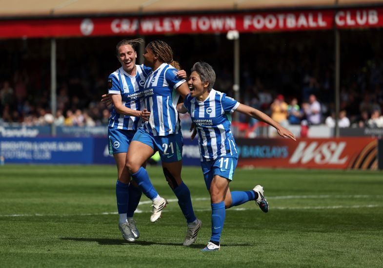 Three players in blue and white striped kits celebrate a goal on a green football pitch at Crawley Town FC with fans in the stands behind them