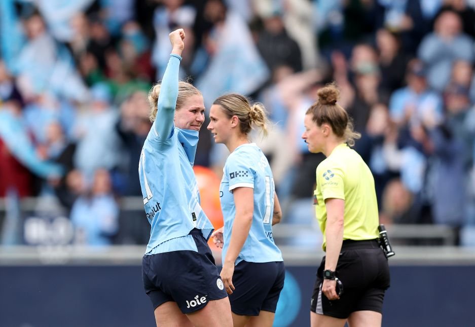 Two female soccer players in light blue jerseys celebrate on the field one with a raised fist as a referee watches in the background