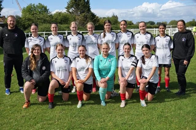 Girls soccer team posing for a group photo on a grassy field wearing white jerseys and red socks with coaches at each end on a sunny day