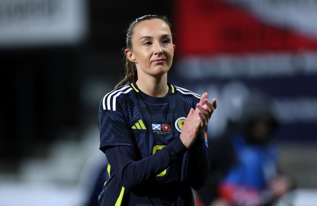 Female soccer player in a dark jersey clapping on the field with national badges Scottish and Swiss flags on the chest