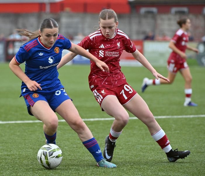 Two female soccer players in blue and red kits battle for the ball on a green field during a match.