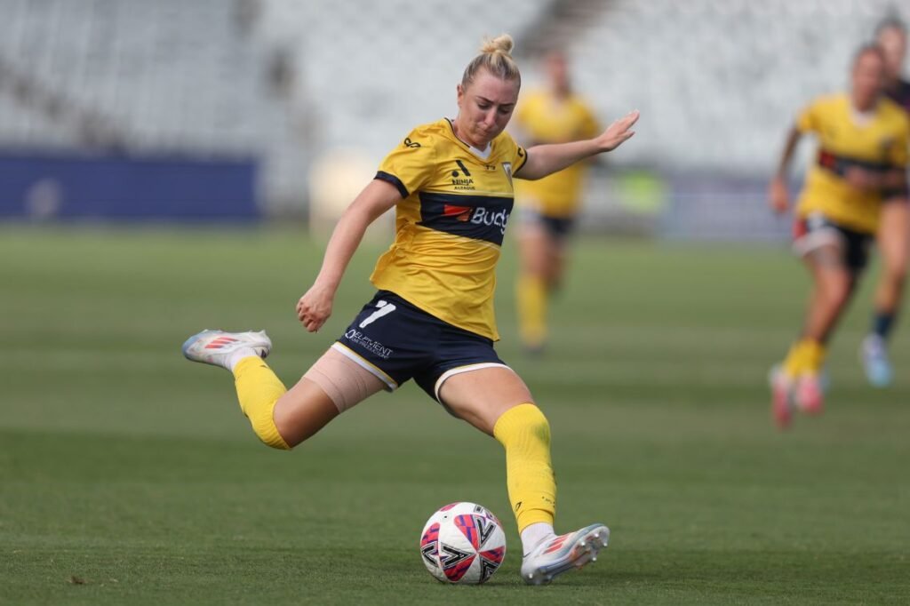 Female soccer player in a yellow kit kicking a ball on a grass field during a match