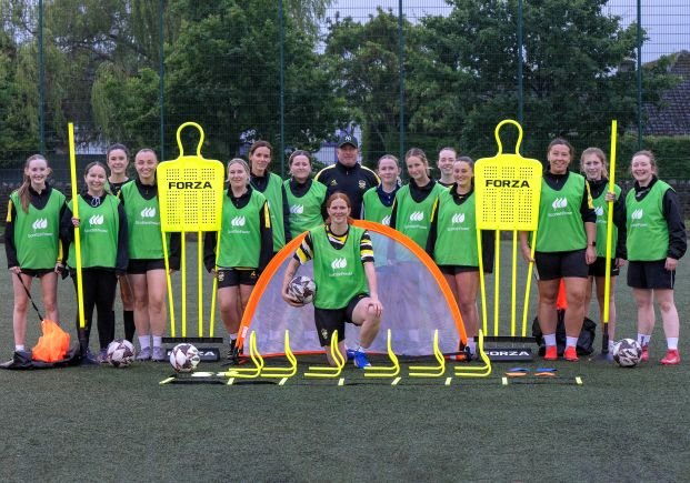 Group of young female soccer players in green bibs posing with coach on a turf field, training equipment arranged in front (cones, ladders, mannequins).