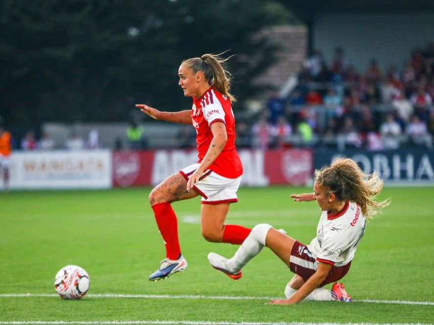 Female soccer players in action a red jersey player runs with the ball while a white jersey opponent slides in from the right ball at their feet on the grass