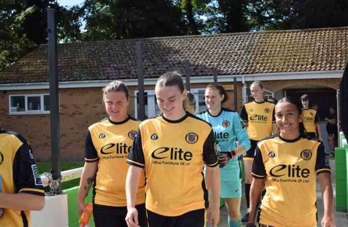 Group of female football players in yellow and black jerseys walking onto the field including a goalkeeper in light blue In a sunny training ground setting