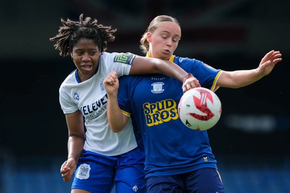 Two female soccer players challenge for the ball during a match one in a white jersey and blue shorts and the other in a blue kit both reaching for the ball close to each other