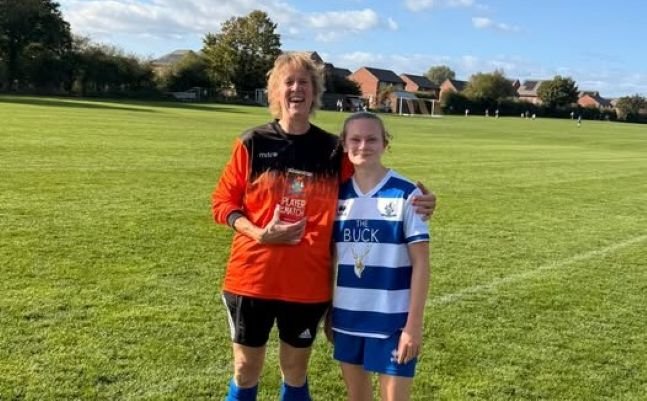 Two smiling soccer players stand on a sunny grassy field; an older woman in orange goalkeeper kit holds a trophy beside a younger girl in blue-and-white stripes.