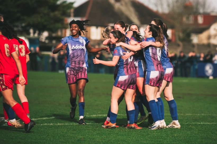 Female soccer players in purple uniforms hug and celebrate on a grassy field after the match