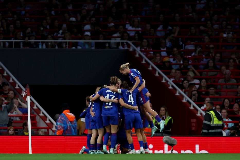 A group of blue clad soccer players huddle and leap in celebration on the pitch as teammates embrace amid a crowded stadium
