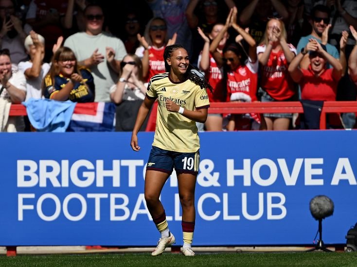 Female soccer player in a pale yellow away kit with number 19 celebrates on the field as cheering fans look on behind a Brighton  Hove Albion banner