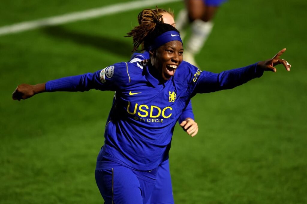 Chelsea women's player in a blue uniform celebrates with arms outstretched on a green soccer pitch after a goal or win.