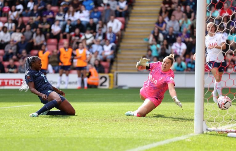 Soccer goalkeeper in pink dives to stop a shot at the goal as a defender slides on the grass nearby